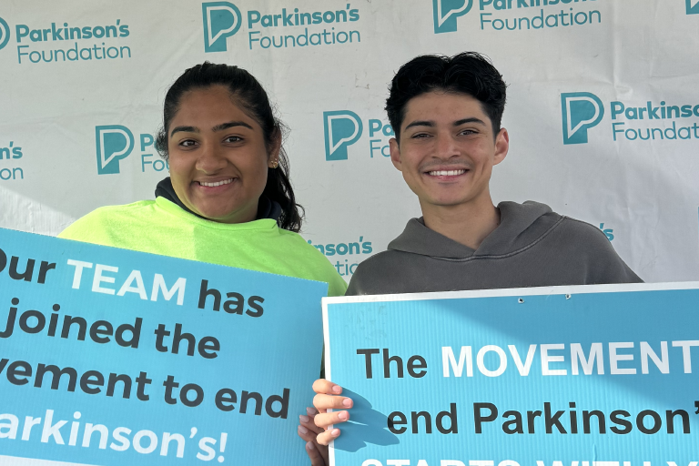 Maya Singh and Nathan Perez standing together and smiling. They are holding posters for the Parkinson's Moving Day event 2025.